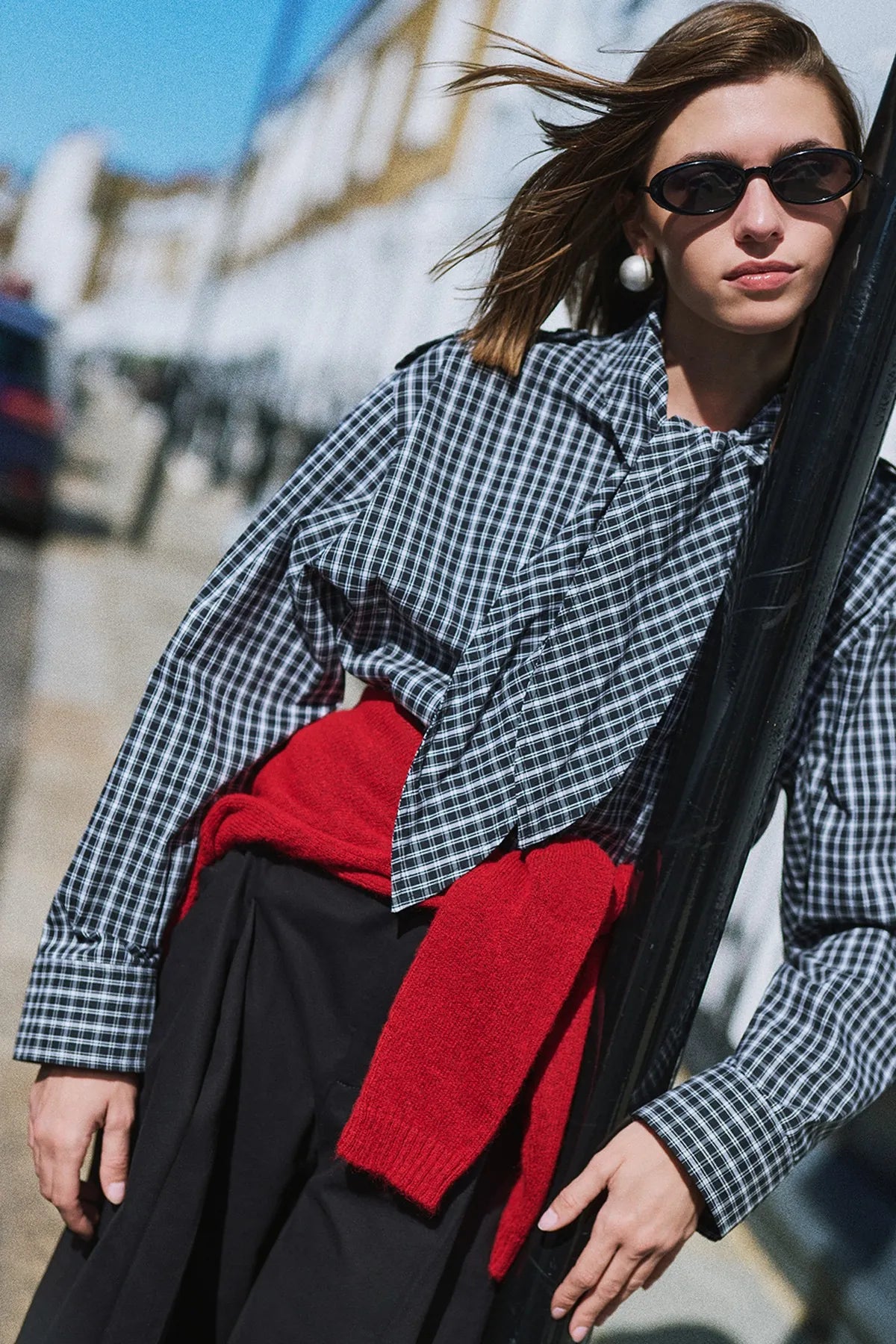 A gingham shirt with a red knit tied over a long black skirt, against a pole.